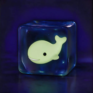 Frostglow Whale glow-in-the-dark cube squishy, clear ice cube toy with luminous pale yellow whale inside on dark blue background