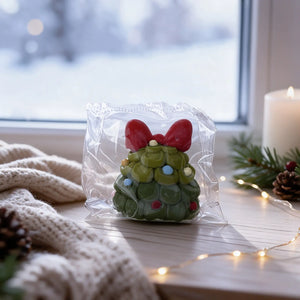 Green Christmas tree-shaped squishy charm with a red bow shown in a wintery setting.