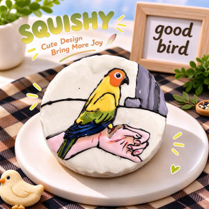 Parrot squishy product photo, round white squishy with hand-painted colorful parrot perched on a hand, bright orange head, yellow body, green wings and blue tail, pet dessert-style scene, brown beige checkered tablecloth, wooden "good bird" sign, bird seed and green plant decorations