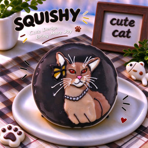 Hand-painted Abyssinian squishy toy, cute Abyssinian cat portrait with pearl necklace and yellow bow on soft round fidget toy, dark glossy surface on display plate