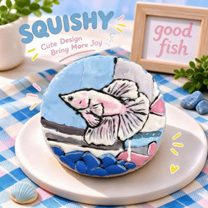 Fish squishy product photo, round blue and white squishy with hand-painted pink betta fish on aquarium glass and stone scene, cute aquatic dessert-style product photo, blue pink checkered tablecloth, wooden "good fish" sign, seashell and blue pebble decorations, bright playful styling