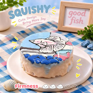 Fish squishy side view, round blue and white squishy with hand-painted pink betta fish, aquarium stones and water detail, soft glossy silicone texture, thick cake-like side profile, blue pink checkered table setting, wooden "good fish" sign in background